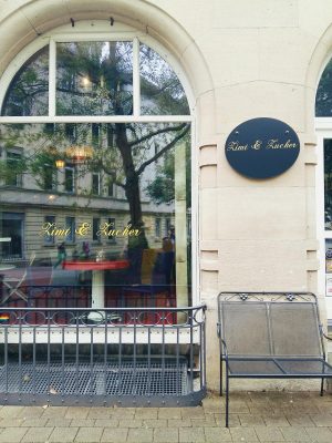 Café mit dem Namen "Zimt und Zucker" mit hohen Fensterbögen. Davor steht eine kleine Metallbank und ein dazu passendes Geländer umgibt den Lüftungsschacht.